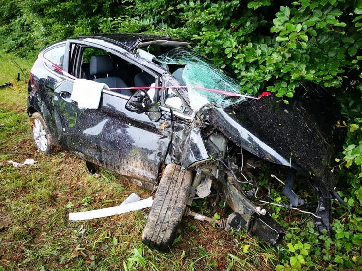 A black car which has crashed into a hedge. The windscreen and front of vehicle is smashed, and the front wheel has come off.